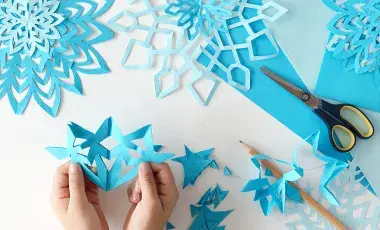 Childs hands holding a blue paper cutout of a snowflake next to a pencil and scissors, and other blue snowflakes.