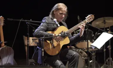 Fabio Zini is shown mid-performance playing a nylon string guitar wearing a shiny black suit. 