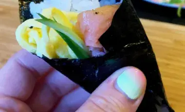 A hand holding a cone shaped roll of seaweed filled with rice and fresh fillings