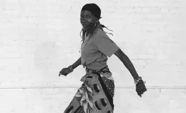 black and white photo of a Black woman standing in a dance pose wearing a boldly printed cloth skirt with a solid t-shirt