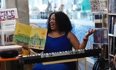 A musician belts out a song while reading a storybook to children. She sits at a musical keyboard and is surrounded by books.