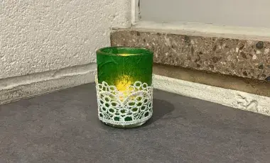 A green votive candle holder wrapped in lace with a lit candle inside