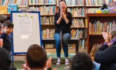 A woman sitsin front of a group of toddlers and adults clapping her hands. A white board is next to her facing the crowd. 