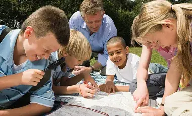 A group of children and an adult are gathered outdoors, examining a map. They look engaged and curious, suggesting a fun learning experience.