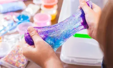 A child stretches a glittery purple and blue slime with both hands. In the background, colorful containers of craft materials are visible on a table. The scene is playful and creative.