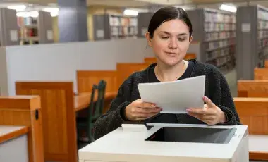A woman gathers pieces of paper from a laser printer in a library.