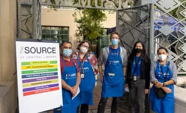 library staff wearing aprons stand in front of resource list sign at the source