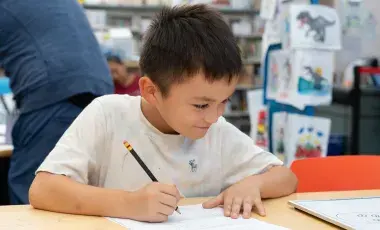A child does homework on a table with other people and bookshelves in the background.