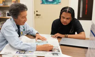 A person helping another person read a newspaper.