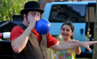 Magician Amazing Dave blowing up a balloon as a smiling child looks on.