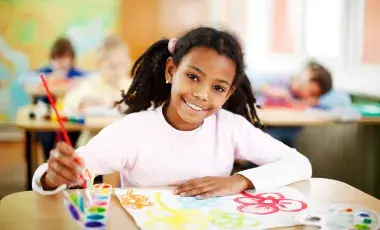 A smiling, young girl uses a paintbrush and colored paint pots to create a watercolor illustration of flowers.