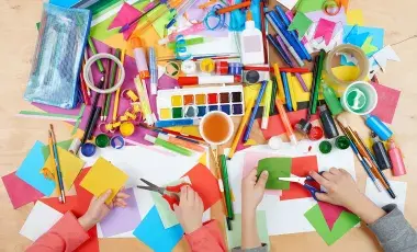 An assortment of craft supplies, paints, markers and hands with scissors cutting paper.
