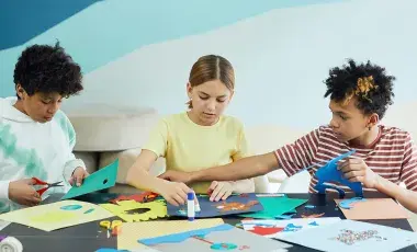 Three children concentrate on making handmade crafts with an assortment of craft supplies.