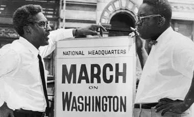 Bayard Rustin at a sign about the March on Washington talking with another man.