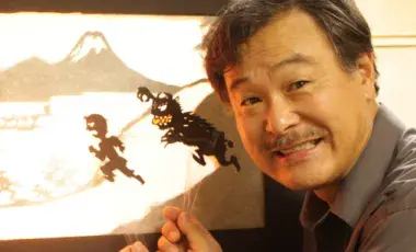 A man is smiling and holding a shadow puppet in front of a screen