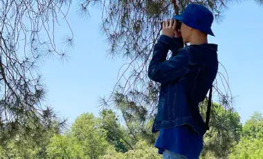 A young man stands under a tree, holding up a pair of binoculars.
