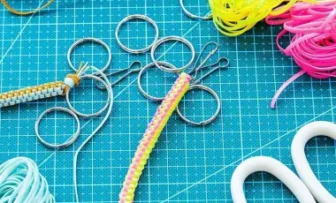 ​The image shows a boondoggle keychain surrounded by all the craft supplies used to make it such as multi-colored gimp string, scissors, and scattered key rings. 