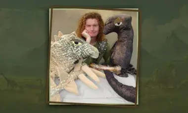 Man with red hair sitting between two dinosaur puppets. 