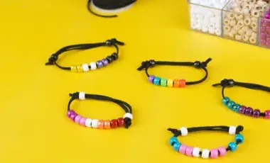 Colorful beaded bracelets on black cords are arranged on a vibrant yellow surface. A box of assorted beads is partially visible in the background.