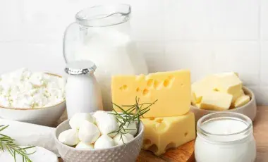Assorted dairy products on a wooden surface, including milk in a jug, various cheeses, yogurt, and cream with sprigs of rosemary, against a tiled background.