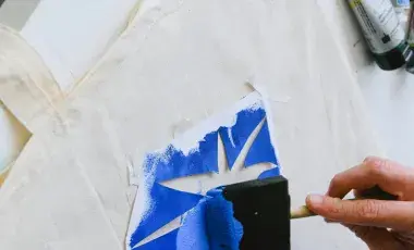 Hand brushing a sponge brush with blue paint over a pointed stencil shape on a natural colored tote bag on a white table.