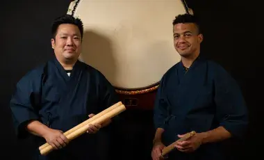 Two BIPOC men in blue wrap coats, holding drumsticks, standing in front of a large taiko drumhead