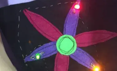 Alternating pieces of magenta and purple petal-shaped felt with a neon green circle piece sewed on a black square of felt with green, pink, and orange LED lights.
