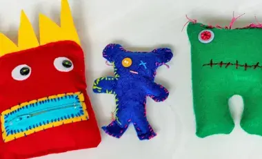 Three hand sewn dolls made of felt against a white background. The doll on the right is a red square with big spiked yellow hair, black button eyes, and a square mouth with yellow lips and light blue zipper The middle doll is dark blue, skinny, and bear-shaped with one button eye on the left and a stitched red X on the right. The doll on the right is a green square with two legs, short pink spikes on it's head, two pink button eyes, and a black uneven line for its mouth