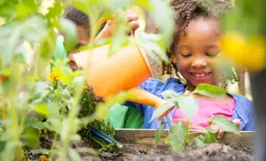 A smiling girl uses a watering can to water garden plants.