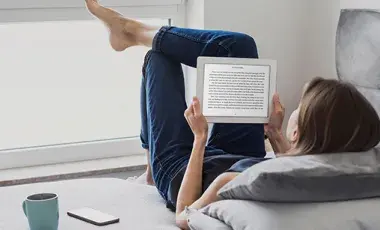 A teenager reclines by a sunny window while reading on her tablet