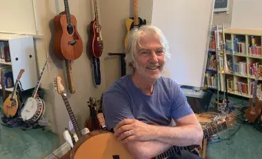 Smiling white-haired white male in blue shirt holding a guitar