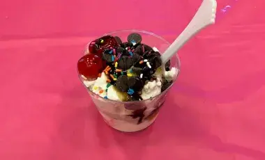 An ice cream sundae topped with cherries, sprinkles, and chocolate chips, on a pink tablecloth