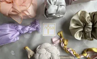 a table decorated with bojagi gift wrappings in various shades of pink, purple, grey, and yellow with a card that says Nossi
