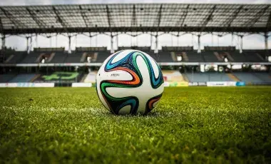 an orange, blue, green, and black wavy design on a white soccer ball which is on top of green grass in a hug stadium