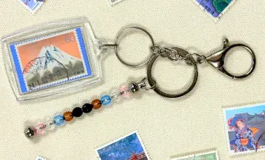 A keychain made of a postage stamp and beads is surrounded by a scattering of postage stamps.