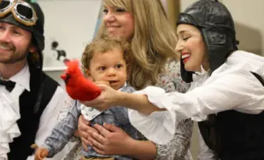 Image is of two LA Opera performers in costume holding a bird prop in front of a young mother with her baby toddler.