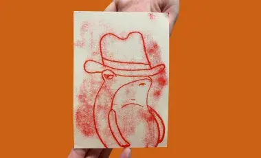 hands holding a mono print of a red frog wearing a cowboy hat