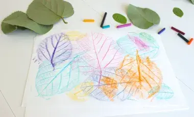 Green leaves are on the table beside a white paper with colorful leaf rubbings featured on the top of the page. 