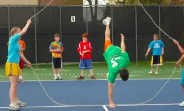 People holding a jump rope on a tennis court, man in center doing a handstand
