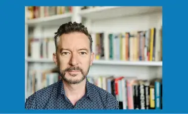 A photograph of Martin Jago wearing a dark blue collared shirt pictured in front of a bookcase.