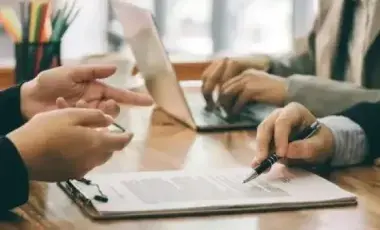 The hands of 3 people at a table signing a document and typing on a laptop