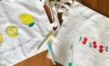 White canvas tote bags decorated with colorful fruit drawings and fabric markers on a table.