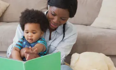 A mother reads from a book to the baby sitting in her lap.