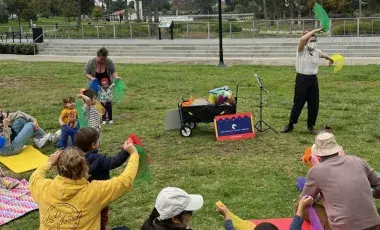 masked librarian leads group of toddlers and caregivers seated on a grassy lawn in singing and waving scarves