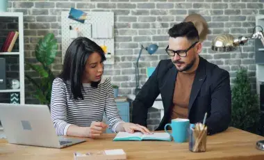 A man and woman sitting at a desk with work materials, looking at a document together