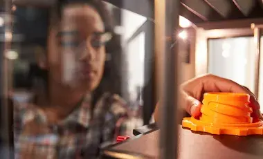 A hand reaching into a 3D printer to touch a 3D printed object on the print bed. 