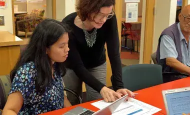 The tutor is standing and pointing to the lesson instructing the learner.