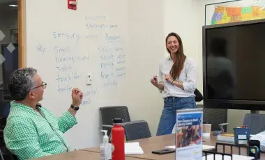 teacher in white blouse and blue jeans long brown hair teaching a learner with grey hair and a light green dress shirt. 