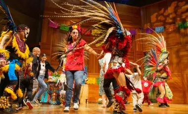 Dancers in complex traditional dress, featuring long feather plumes and colorful clothing, dance on a stage