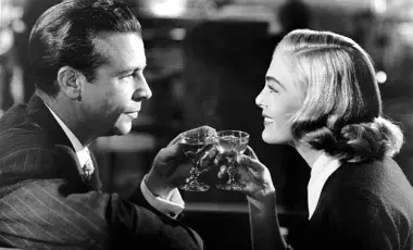 The protagonists, a man and a woman, are looking at each other smiling and holding glasses with alcoholic drinks